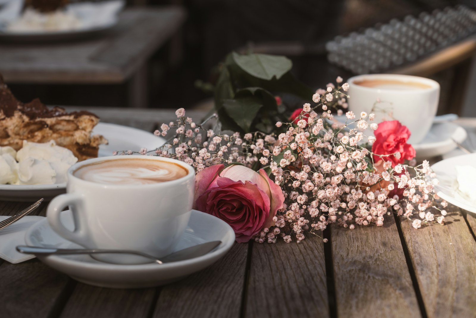 Two coffees beside fresh roses and a slice of cake on a wooden table